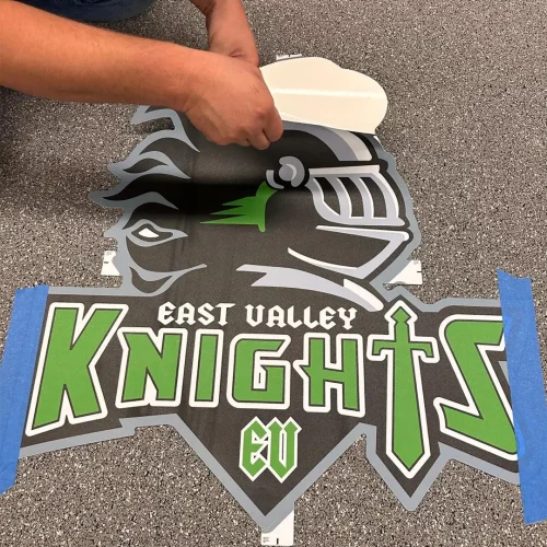 Installer applying an East Valley Knights engineered floor logo onto Steel Appeal performance rubber flooring, peeling back the protective adhesive layer.