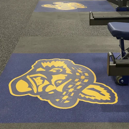 Close-up of two engineered floor graphics installed on blue inlaid rubber platforms with gray drop zones, surrounded by dark gray rubber flooring with large white and black granules. A yellow-and-blue Scottish warrior graphic appears in front, and a yellow-and-blue HP monogram is behind it, with part of the bench and rack safety bars visible.