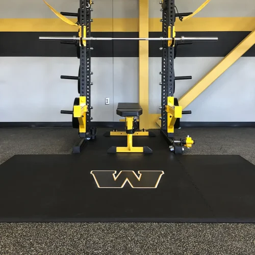 Engineered “W” floor logo installed on an American Platforms rubber weightlifting platform, with a black rack featuring yellow J-hooks and yellow safety bars.